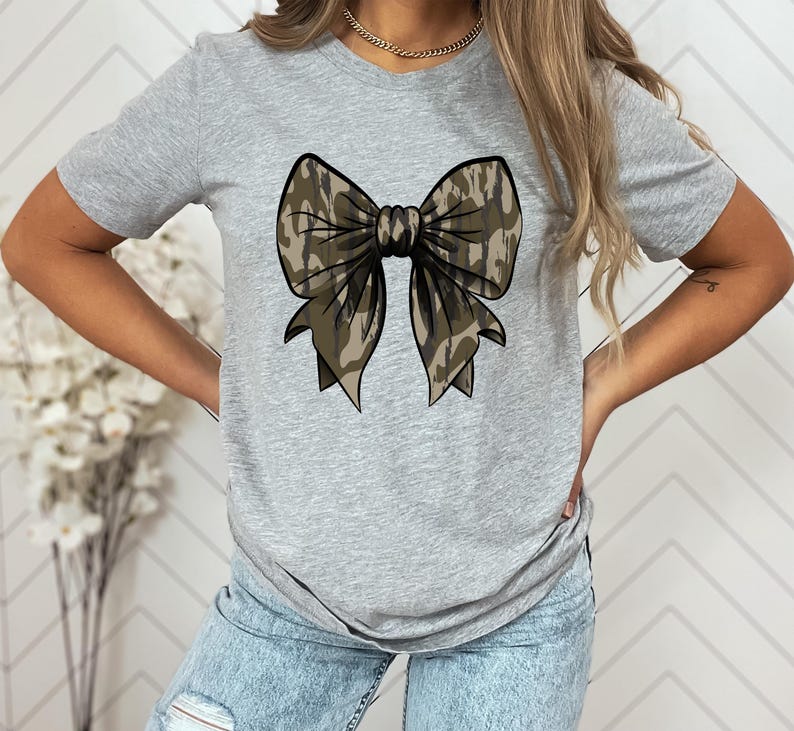 Puede incluir: Una camiseta gris con un gran lazo de camuflaje estampado en la parte delantera.