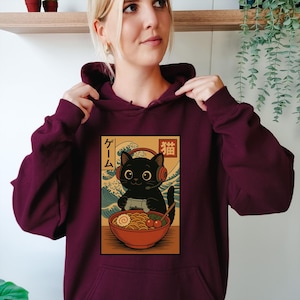 May include: Burgundy hoodie featuring a graphic of a black cat wearing headphones and holding a game controller, with a bowl of ramen. Japanese text is included in the design. The cat is set against a backdrop of ocean waves.