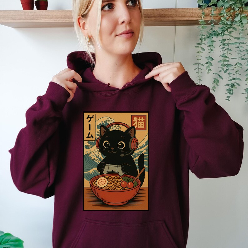 May include: Burgundy hoodie featuring a graphic of a black cat wearing headphones and holding a game controller, with a bowl of ramen. Japanese text is included in the design. The cat is set against a backdrop of ocean waves.