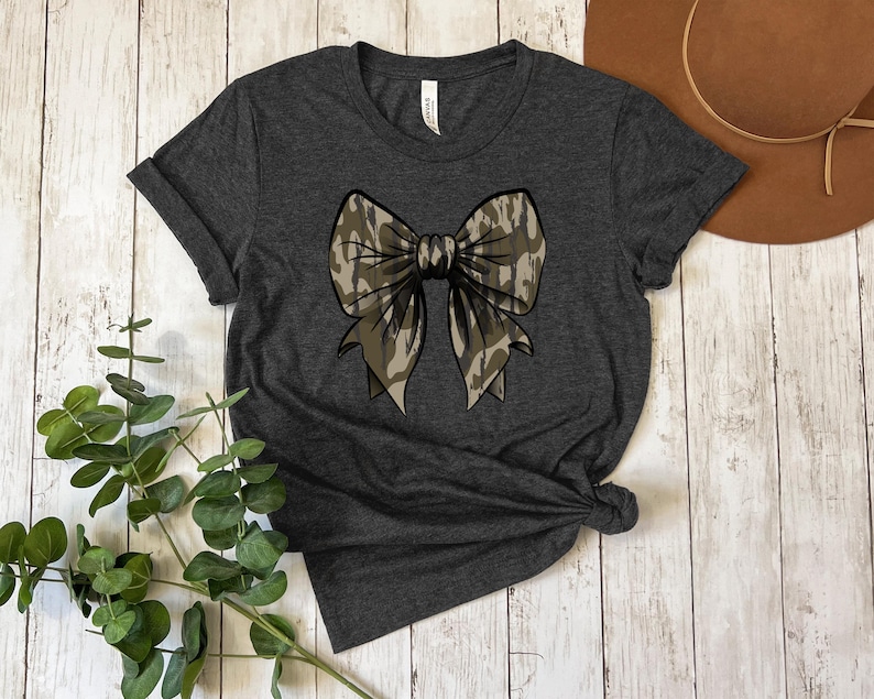 Puede incluir: Una camiseta gris oscuro con un gran lazo de camuflaje estampado en la parte delantera.