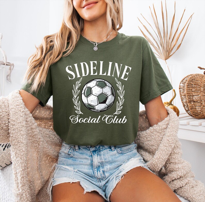 Puede incluir: Camiseta verde oliva con el texto blanco "SIDELINE Social Club". Un gr&aacute;fico de bal&oacute;n de f&uacute;tbol est&aacute; centrado, enmarcado por una corona de laurel. La camiseta es de estilo casual.