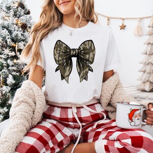 Puede incluir: Una camiseta blanca con un gran dise&ntilde;o de lazo de camuflaje. La persona lleva pantalones de pijama a cuadros rojos y blancos.