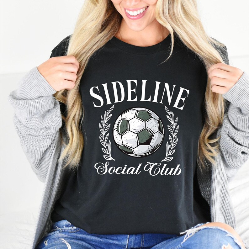 Puede incluir: Camiseta negra con las palabras "SIDELINE Social Club" en blanco, con un gr&aacute;fico de bal&oacute;n de f&uacute;tbol. El dise&ntilde;o incluye un bal&oacute;n de f&uacute;tbol en el centro, rodeado por una corona de laurel. La camiseta es de estilo casual.