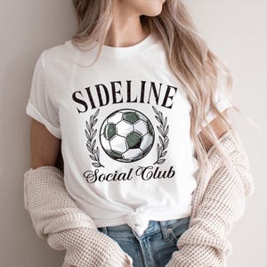 White shirt with “Sideline Social Club” text surrounding a soccer ball framed by laurel leaves. Retro sports design for moms, fans, and soccer families.