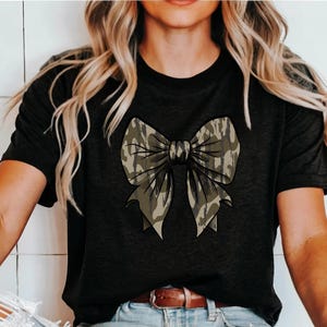 Puede incluir: Una camiseta negra con un gran lazo de camuflaje en la parte delantera. El lazo es en tonos de verde y marr&oacute;n.