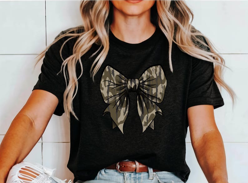 Puede incluir: Una camiseta negra con un gran lazo de camuflaje en la parte delantera. El lazo es en tonos de verde y marr&oacute;n.