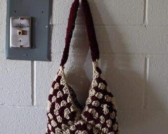 Puede incluir: Un bolso de crochet hecho a mano con un patrón a cuadros burdeos y crema cuelga de un gancho blanco. El bolso tiene forma redondeada y una correa burdeos. El fondo es una pared de ladrillos blancos con una caja eléctrica gris.