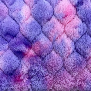 May include: A close-up of a plush, textured fabric in a diamond pattern. The fabric is a blend of purple, pink, and lavender hues, creating a soft, fluffy appearance. The design is reminiscent of a cozy blanket or rug.