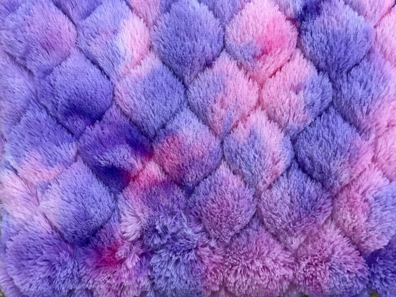 May include: A close-up of a plush, textured fabric in a diamond pattern. The fabric is a blend of purple, pink, and lavender hues, creating a soft, fluffy appearance. The design is reminiscent of a cozy blanket or rug.