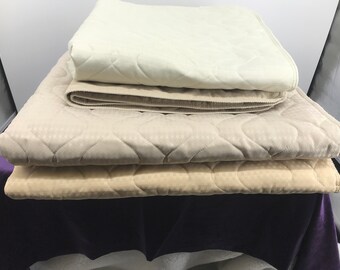 May include: Three quilted bed pads in different shades of beige, stacked on top of each other. The top pad is a lighter shade of beige, the middle pad is a medium shade of beige, and the bottom pad is a darker shade of beige.