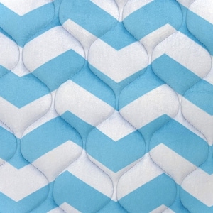 Puede incluir: Primer plano de una tela acolchada con un patrón de chevrón repetido. El diseño presenta rayas alternas en azul claro y blanco, creando una apariencia texturizada y acolchada. Las costuras siguen las curvas del patrón.