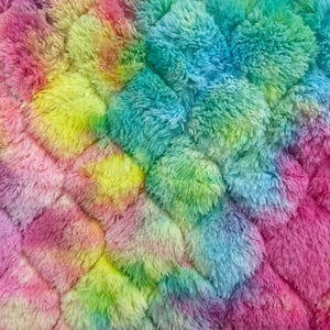 May include: Close-up of a soft, textured fabric in a vibrant rainbow of colors. The plush material features a repeating pattern of diamond-shaped tufts, with hues of pink, yellow, green, and blue blending together. The overall effect is a colorful and cozy textile.