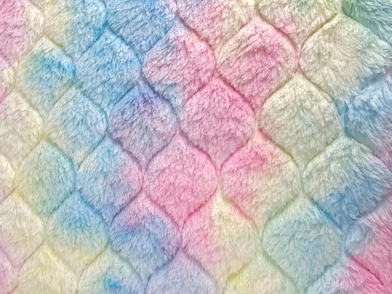 May include: Close-up of a soft, plush fabric with a mermaid scale pattern. The fabric features a gradient of colors, including pink, blue, yellow, and white. The texture is fluffy and inviting, creating a whimsical and cozy feel.