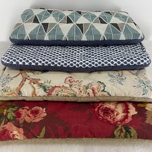 May include: Four rectangular pet beds stacked on top of each other. The top bed has a geometric pattern in shades of blue, gray, and white. The second bed has a blue dotted pattern. The third bed has a floral pattern. The bottom bed has a red floral pattern.
