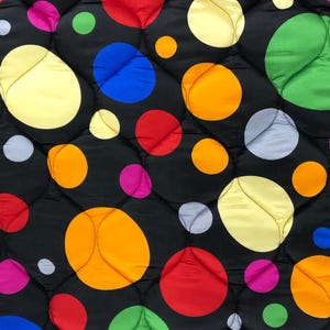 May include: A close-up of a quilted fabric featuring a black background and large, colourful polka dots. The dots are in shades of yellow, orange, red, pink, blue, green, and grey, arranged in a repeating pattern.