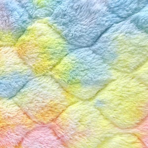 May include: Close-up of a soft, plush fabric with a quilted pattern. The material is a vibrant blend of colors, including blue, yellow, orange, and pink, creating a rainbow effect. The texture appears fluffy and inviting.