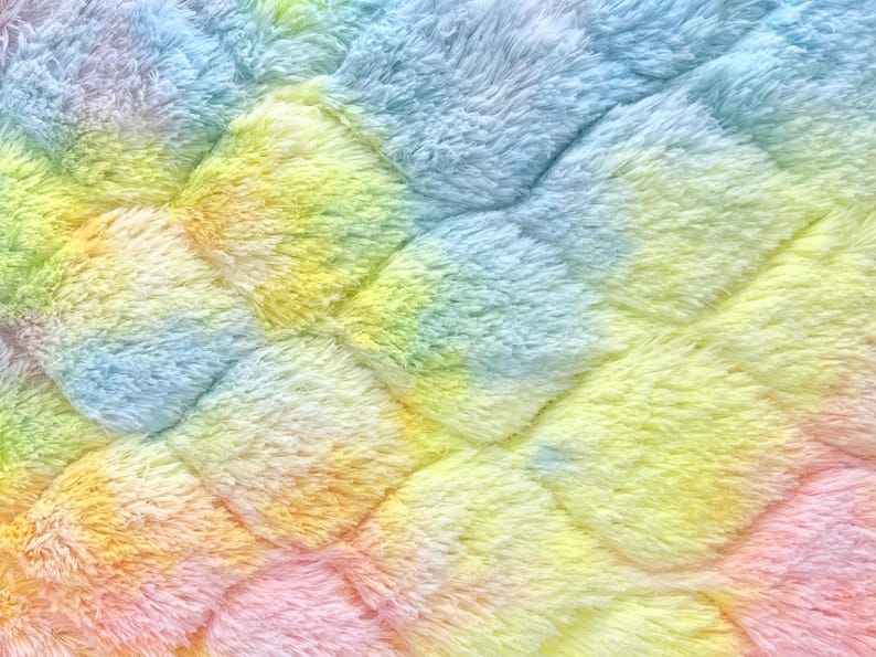 May include: Close-up of a soft, plush fabric with a quilted pattern. The material is a vibrant blend of colors, including blue, yellow, orange, and pink, creating a rainbow effect. The texture appears fluffy and inviting.