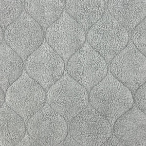 Puede incluir: Una tela de color gris claro con una textura afelpada y suave, que presenta un patrón acolchado repetitivo de formas ojivales entrelazadas. La superficie tiene un aspecto táctil y mullido con líneas de costura que definen el diseño.
