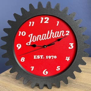 May include: A black gear-shaped clock with a red face. The clock face features white numerals, the name "Jonathan" in cursive, and the text "EST. 1970". The clock is displayed on a wooden surface.