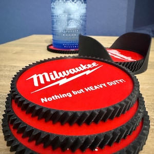 May include: A stack of three circular red coasters with black gear-shaped edges. Each coaster features the white Milwaukee logo and the text Nothing but HEAVY DUTY! A black holder contains additional coasters, and a blue glass sits on one coaster.
