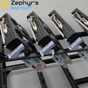May include: A black metal mining rig frame holding multiple Sapphire graphics cards. Each card is secured with light grey 3D-printed plastic brackets attached to the frame. The cards feature visible cooling fans, metallic heat pipes, and ports.