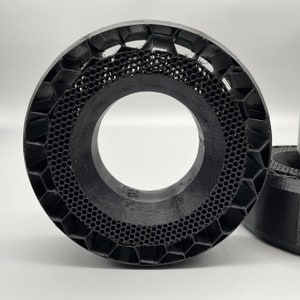 Puede incluir: Un anillo negro impreso en 3D con un patr&oacute;n de panal. El anillo tiene una gran abertura central y m&uacute;ltiples capas de perforaciones hexagonales y circulares. El dise&ntilde;o es intrincado y geom&eacute;trico, con un enfoque en la textura y la forma.