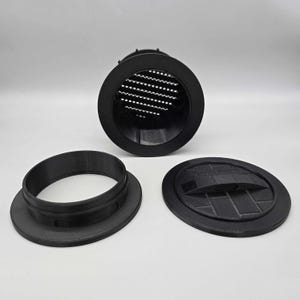 May include: A black circular vent component with a grid pattern, a separate circular mounting ring, and a flat circular cover featuring a central handle and a brick-like embossed pattern on its surface, all constructed from matte black material.