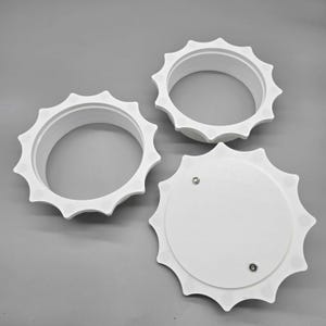May include: Three white circular components with scalloped, gear-like edges. Two of the pieces are open rings with internal threading, while the third piece is a solid circular cover featuring two small metal fasteners on its flat surface.