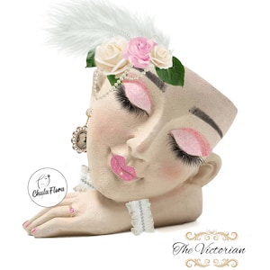 May include: A white ceramic planter shaped like a woman's face with closed eyes, pink lips, and a white feather and pink flower crown. The planter has a pink glitter finish and a white lace trim around the neck.