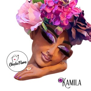May include: A woman with a flower crown on her head, wearing heavy makeup with glitter and rhinestones. She is wearing a ring on her finger and has her arm resting on her chin. The background is white.