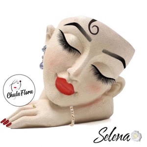 May include: White ceramic planter shaped like a woman's face with closed eyes, red lips, and a gold ring on her finger. The planter has a small chain necklace around her neck. The planter is labeled "Selena" with a flower.