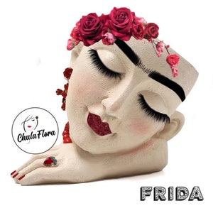 May include: White ceramic planter shaped like Frida Kahlo's face with a red rose flower crown, closed eyes, and red lips. The planter is lying on its side with a hand resting on the surface. The text "FRIDA" is written in silver glitter letters below the planter.