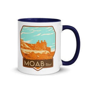 May include: White ceramic coffee mug with a blue rim and handle. The mug features a vintage-style illustration of a red rock formation with the text "MOAB Utah" in brown lettering.