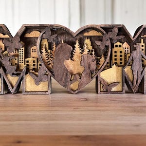 May include: Wooden cutout of the word "MOM" with a heart in the middle. The heart features a silhouette of a couple standing in a forest. The letters are decorated with silhouettes of buildings, trees, and birds.