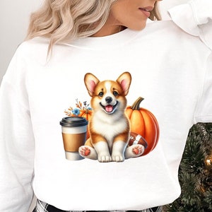 Może przedstawiać: Biała bluza z długim rękawem z grafiką przedstawiającą psa rasy corgi trzymającego piłkę nożną i kubek kawy. Corgi otoczony jest jesiennymi elementami, takimi jak dynie i liście.