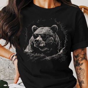 Puede incluir: Camiseta negra con un gráfico de un oso con gafas de sol. El oso está en un entorno de bosque oscuro con un cielo estrellado.