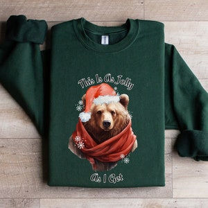 May include: Green Christmas sweatshirt featuring a brown bear wearing a Santa hat and a red scarf. The text on the sweatshirt reads "This is as Jolly as I Get."