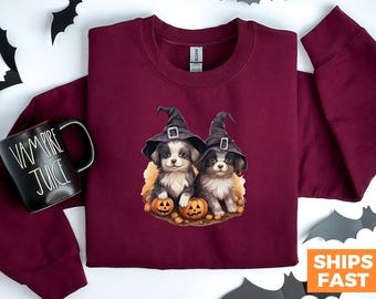 May include: A maroon sweatshirt with a graphic of two dogs wearing witch hats and holding pumpkins. The dogs are brown and white with black noses. The pumpkins are orange and have carved faces.