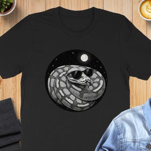 May include: Black t-shirt featuring a circular graphic of a snake wearing sunglasses under a starry night sky with a full moon. The snake is coiled and detailed in black and white. The shirt is displayed on a wooden surface.