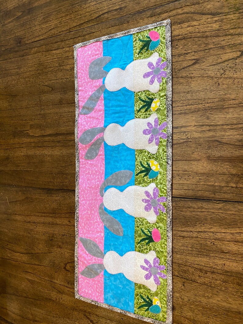 以下が含まれることがあります： ピンク、ブルー、グリーンのストライプの背景を持つキルトのテーブルランナー。グレーの耳を持つ白いウサギが4匹、ランナーに並んでおり、それぞれ紫の花と緑の葉が付いています。