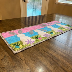 以下が含まれることがあります： 緑、青、ピンクの背景に、灰色の耳を持つ白いウサギが並んだテーブルランナー。ウサギは緑の葉、紫の花、黄色の卵に囲まれています。