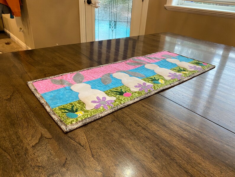 以下が含まれることがあります： 緑、青、ピンクの背景に、灰色の耳を持つ白いウサギが並んだテーブルランナー。ウサギは緑の葉、紫の花、黄色の卵に囲まれています。