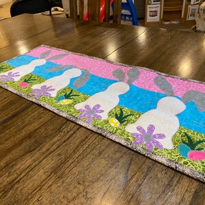 以下が含まれることがあります： ピンク、ブルー、グリーンのパッチワークデザインの白いテーブルランナーで、グレーの耳を持つ白いウサギが5匹描かれています。ウサギは一列に並んでおり、同じ方向を見ています。ランナーはグレーとホワイトのボーダーが付いています。