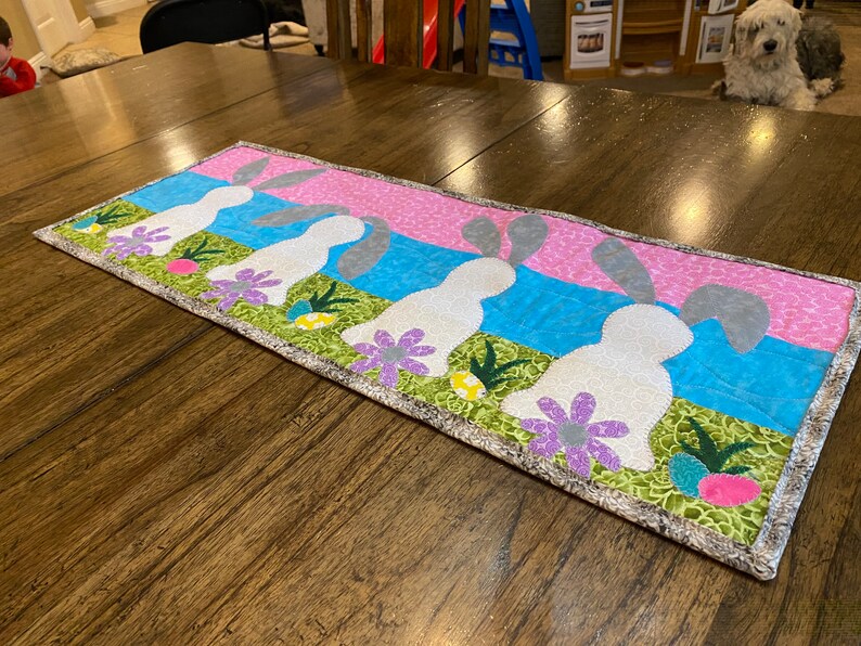 以下が含まれることがあります： ピンク、ブルー、グリーンのパッチワークデザインの白いテーブルランナーで、グレーの耳を持つ白いウサギが5匹描かれています。ウサギは一列に並んでおり、同じ方向を見ています。ランナーはグレーとホワイトのボーダーが付いています。