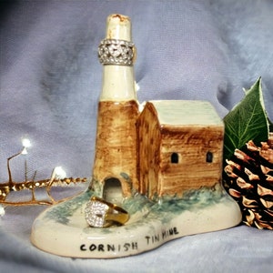 Pode incluir: Um porta-anéis de cerâmica em forma de mina de estanho da Cornualha. O suporte tem um exterior marrom e branco com uma base verde. As palavras "Cornish Tin Mine" estão escritas na base.