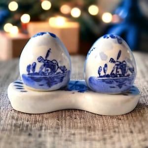 Puede incluir: Un par de saleros y pimenteros de cerámica azul y blanca con un diseño de molino de viento holandés, sentados sobre una base de cerámica blanca.