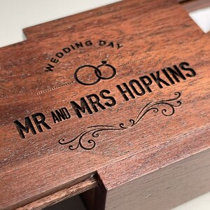 May include: A wooden ring box with an engraved inscription that reads "Wedding Day" and "Mr. and Mrs. Hopkins". The box has a hinged lid and a small compartment for rings.