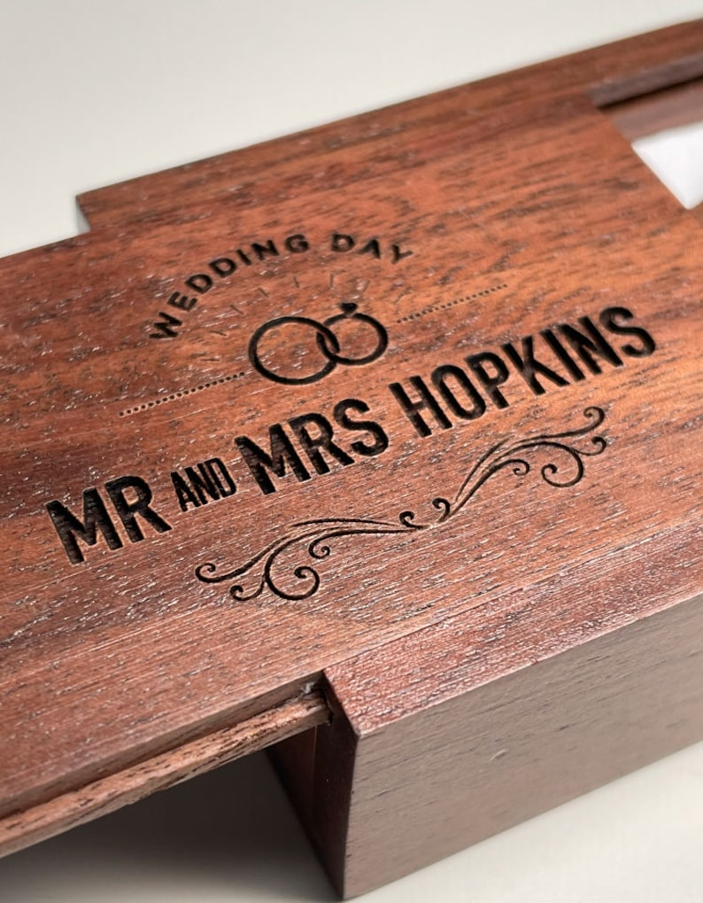 May include: A wooden ring box with an engraved inscription that reads "Wedding Day" and "Mr. and Mrs. Hopkins". The box has a hinged lid and a small compartment for rings.