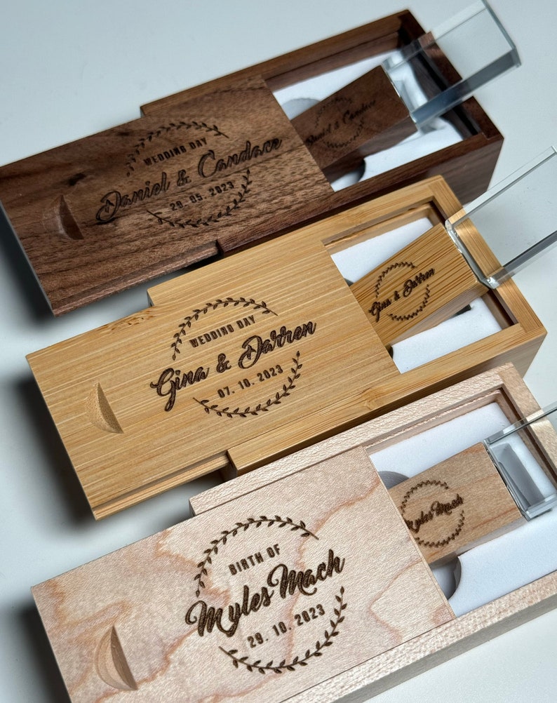 May include: Three wooden boxes with engraved text and clear glass lids. Each box contains a USB drive with a personalized inscription. The inscriptions include "Wedding Day Daniel & Candace 29.05.2023", "Wedding Day Gina & Darren 07.10.2023", and "Birth of Myles Mach 29.10.2023".