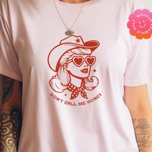 May include: Pink t-shirt with a red graphic of a woman wearing a cowboy hat and heart-shaped sunglasses. The text "Don't Call Me Honey" is printed below the graphic.
